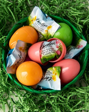 A joyful Easter scene set outdoors with Chocolove chocolate eggs nestled among fresh spring grass. A pastel blue Easter basket, filled with decorated eggs, sits nearby, evoking the excitement of an Easter egg hunt. Soft natural lighting enhances the festive and cheerful atmosphere.