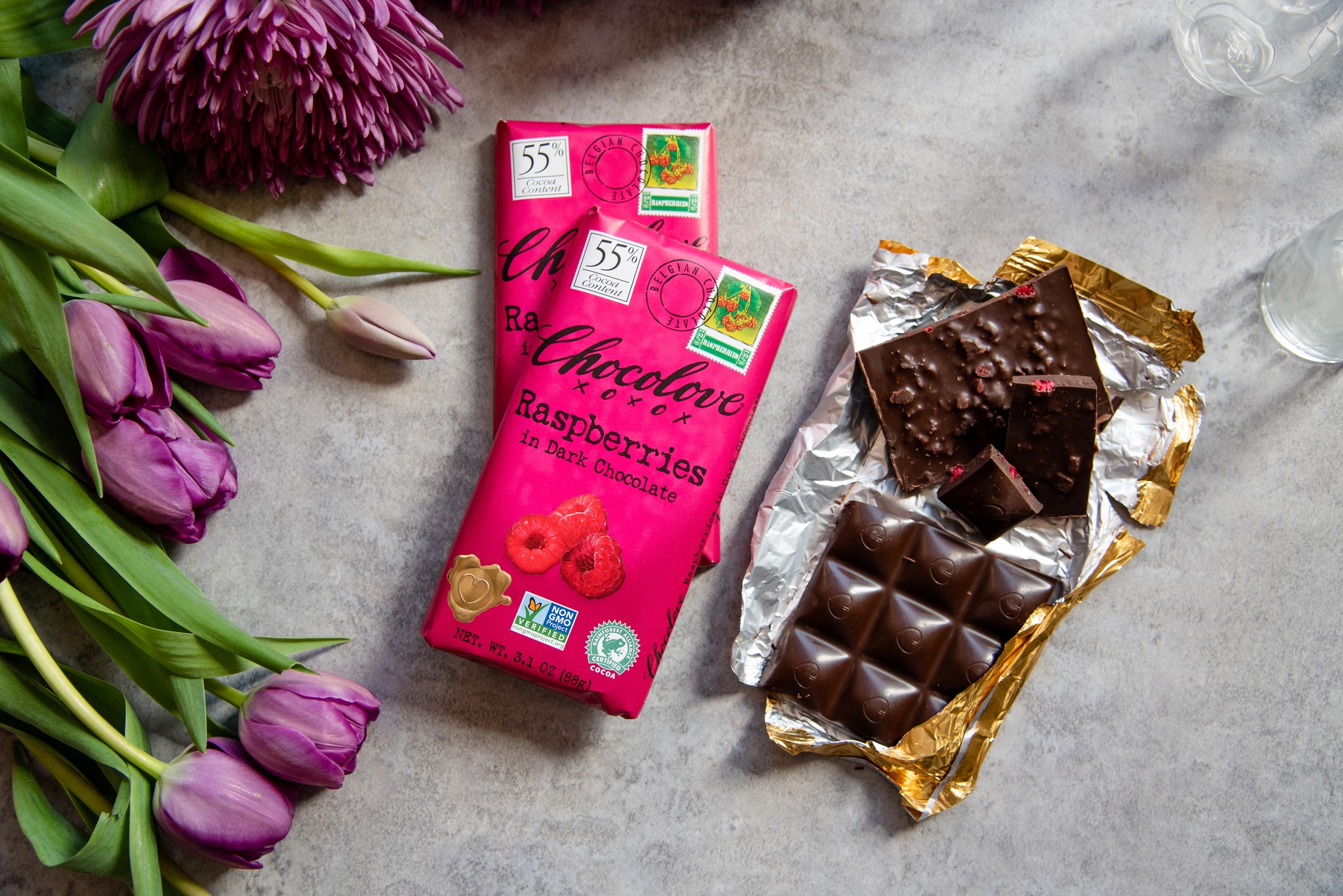 Chocolove Raspberry in Dark Chocolate bars, one opened and still in its foil wrapper, arranged next to pink spring flowers on a grey marble surface