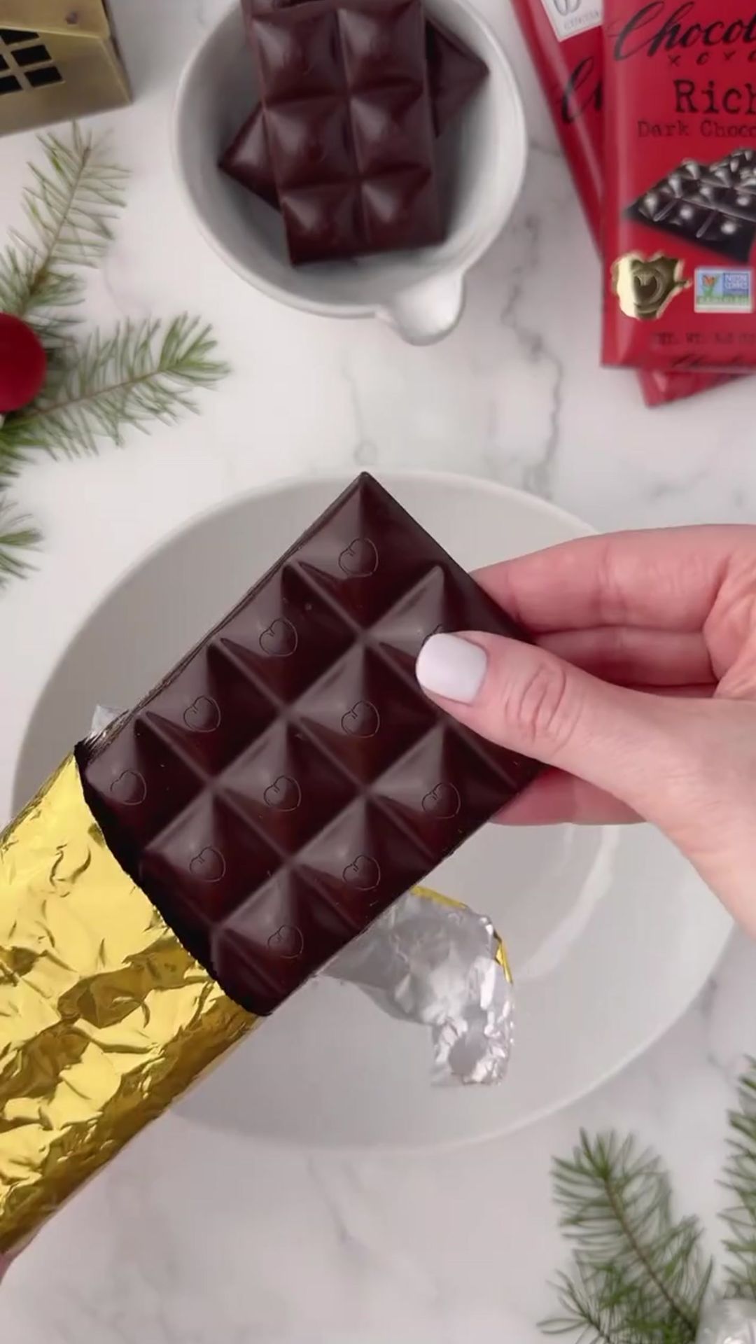 Close-up of a hand holding a Chocolove Rich Dark Chocolate Bar wrapped in golden foil, ready to be unwrapped. The background features holiday-themed decor, including evergreen sprigs, a bowl of chocolate squares, and additional Chocolove chocolate bars, creating a festive atmosphere.
