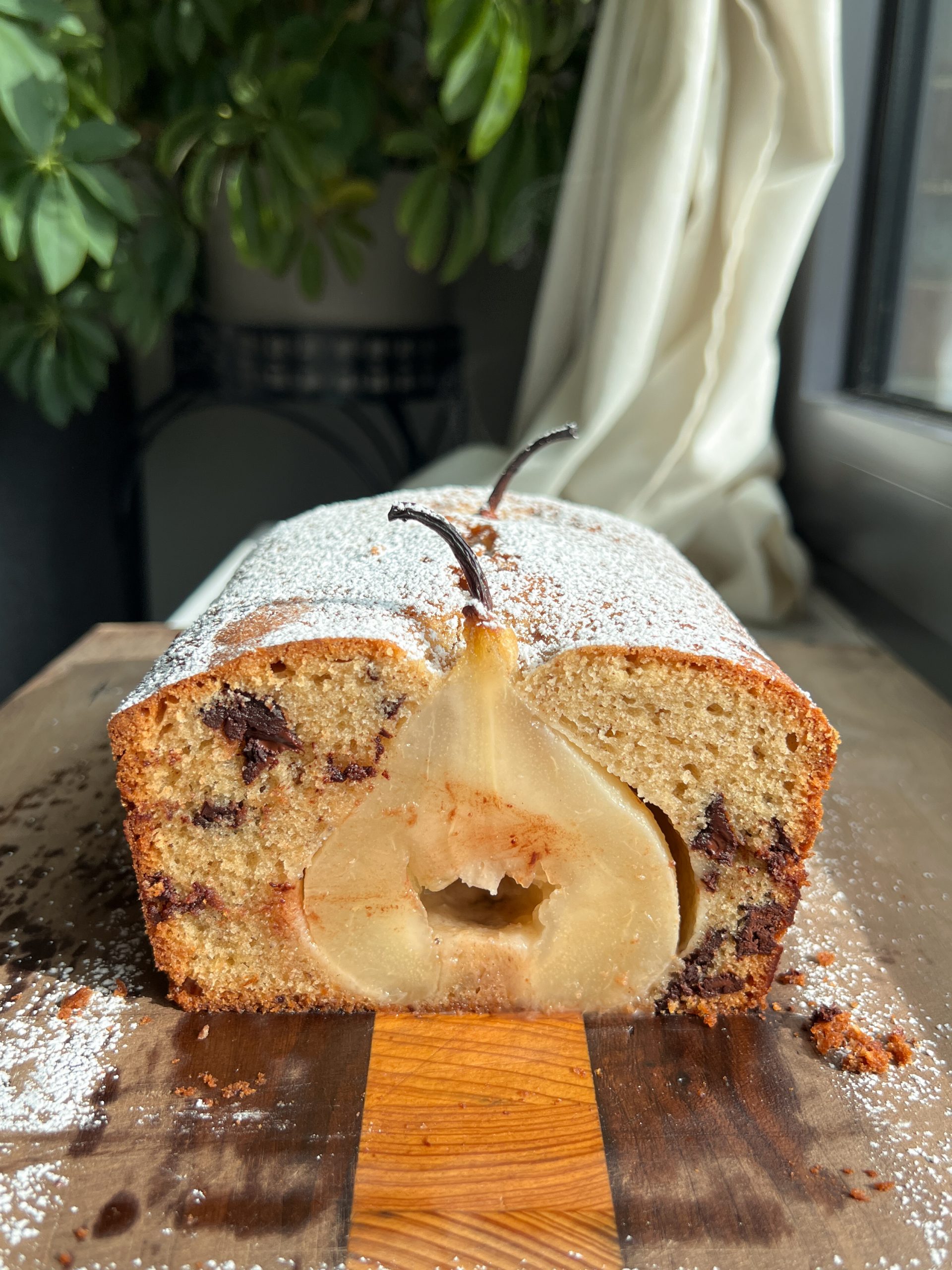 A sliced pear loaf cake showing a whole poached pear baked inside, surrounded by golden crumb and dark chocolate chunks, lightly dusted with powdered sugar.
