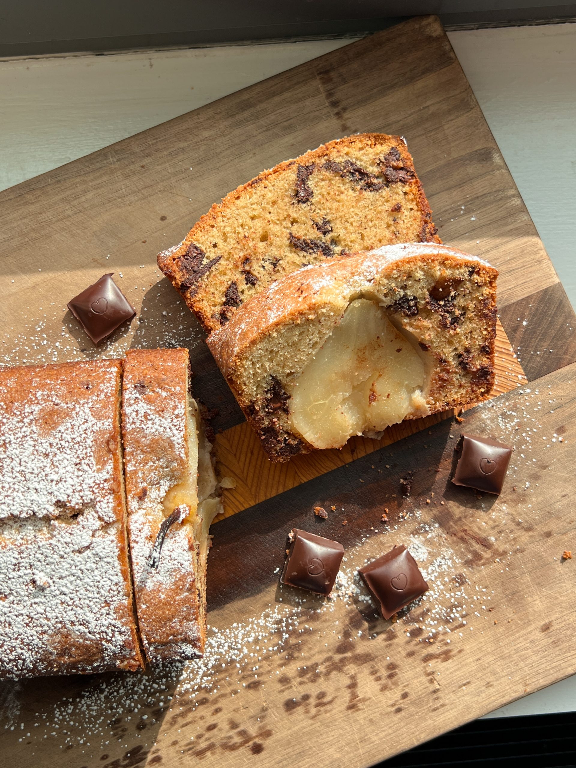 Slices of pear loaf cake with visible dark chocolate chunks arranged on a wooden board, surrounded by Chocolove chocolate squares and dusted with powdered sugar.