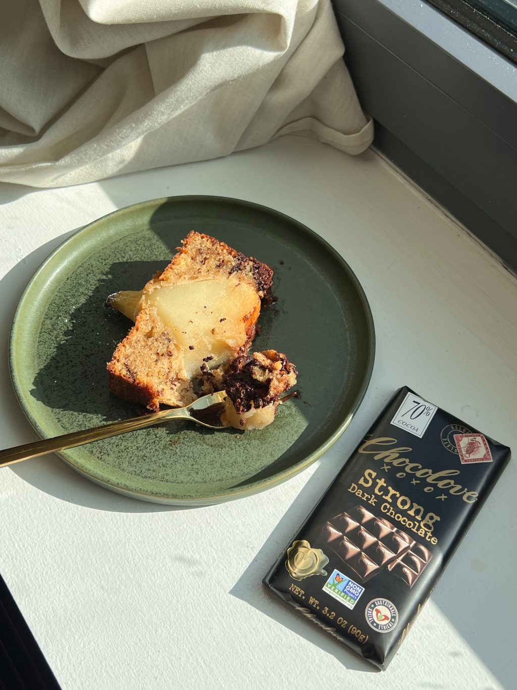 A slice of pear and dark chocolate loaf cake on a green plate beside a bar of Chocolove Strong Dark Chocolate, captured in warm natural light.