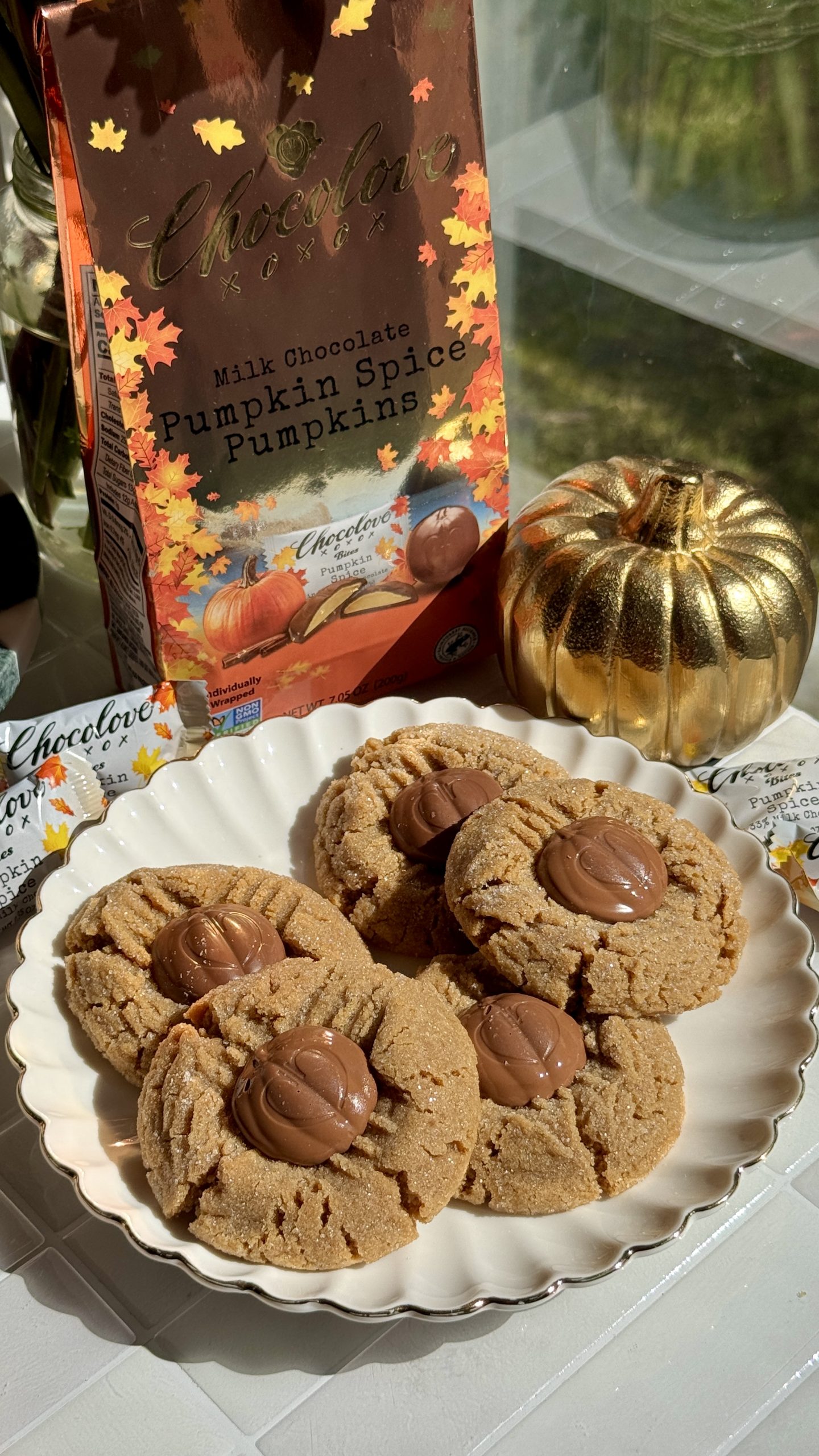 Plate of peanut butter cookies topped with Chocolove Milk Chocolate Pumpkin Spice Pumpkins next to fall-themed Chocolove packaging and golden pumpkin décor in natural sunlight.